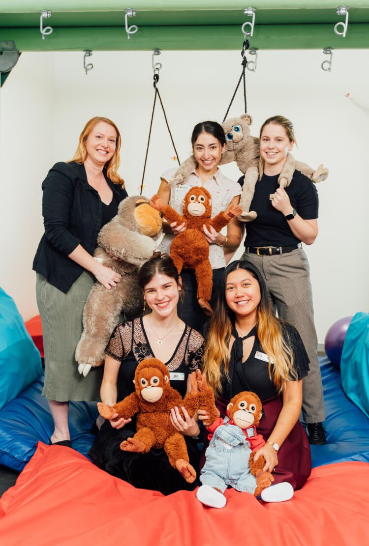 A group of five people pose indoors, holding plush toys and wearing name tags. They are surrounded by colorful cushions and playful, hanging decorations.