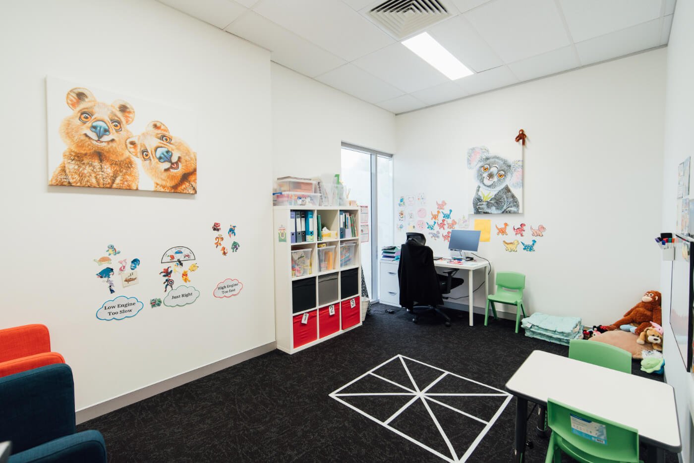 A bright, tidy children's therapy room with wall art, a cubby of toys, a desk with a computer, and child-sized tables and chairs.