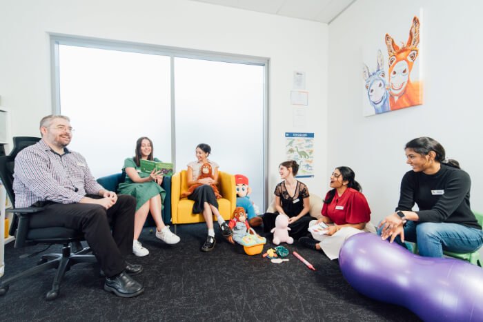 Pepper Kids Therapy team are sitting in a brightly lit room with toys around them, engaging in conversation. Some are sitting in chairs while others are on the floor.