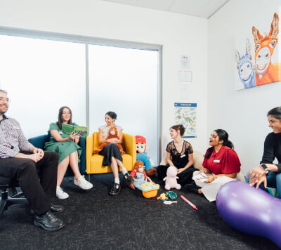 A group of people sitting in a room, some in colorful chairs, engaging in conversation. Toys are scattered on the floor, and a bright painting of llamas hangs on the wall.