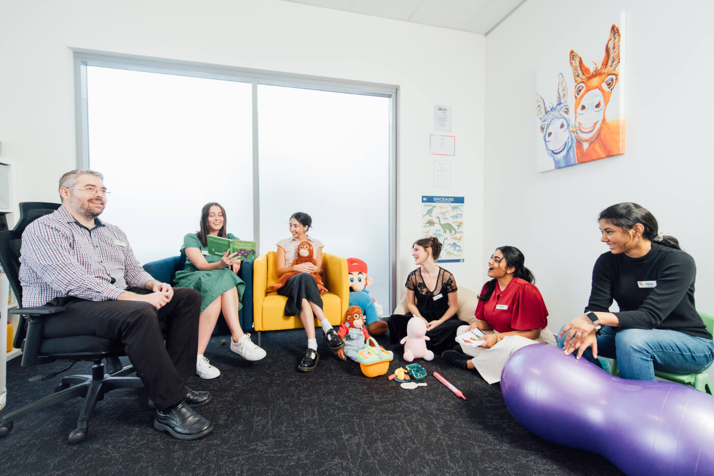 A group of six people are seated in a circle in a brightly lit room, engaging in a lively conversation. The room contains colorful toys and a wall with artwork depicting two animal-like figures.
