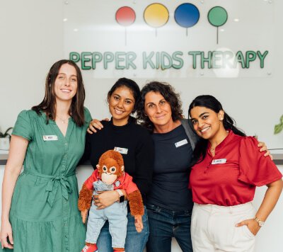 Four women stand together smiling in front of a sign that reads "Pepper Kids Therapy." One woman holds a stuffed monkey.