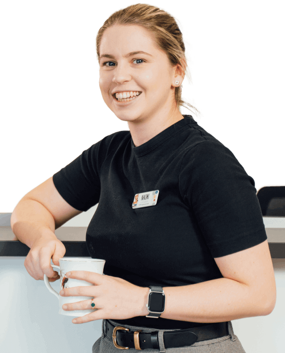 A person wearing a black shirt, gray pants, and a name badge is holding and stirring a mug. They are smiling and standing in front of a white background.